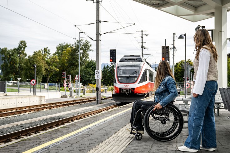 Frau mit Rollstuhl und stehende Frau warten am Bahnsteig auf einfahrenden Zug