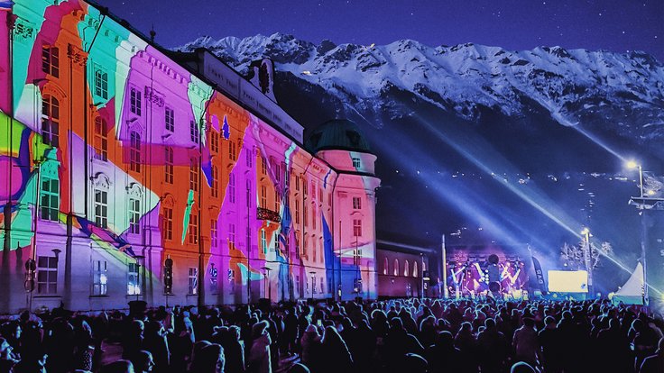 Bergsilvester Konzert in der Nacht mit vielen Menschen vor Bühne neben bunt beleuchteter Hofburg und Nordkette im Hintergrund mit Schnee auf Gipfeln.