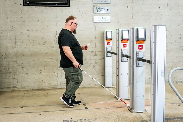 Mann mit Blindenstock vor Zutrittssytem/Ticketschranken