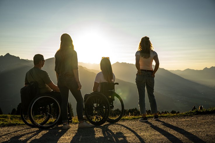 Zei Rollstuhlfahrer und zwei stehende Personen am Berg bei Sonnenuntergang